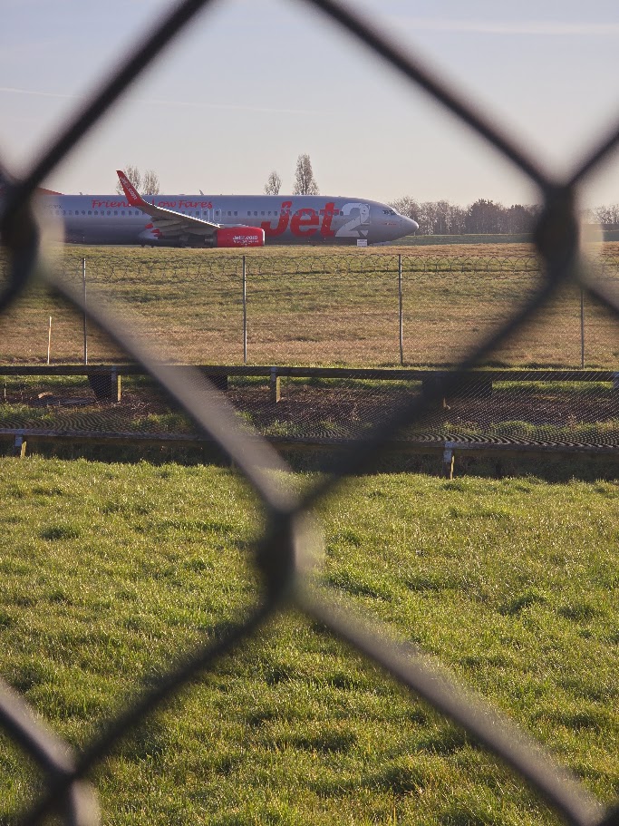 Jet2 Boeing 737 taxiing at Birmingham Airport, viewed through the perimeter fence