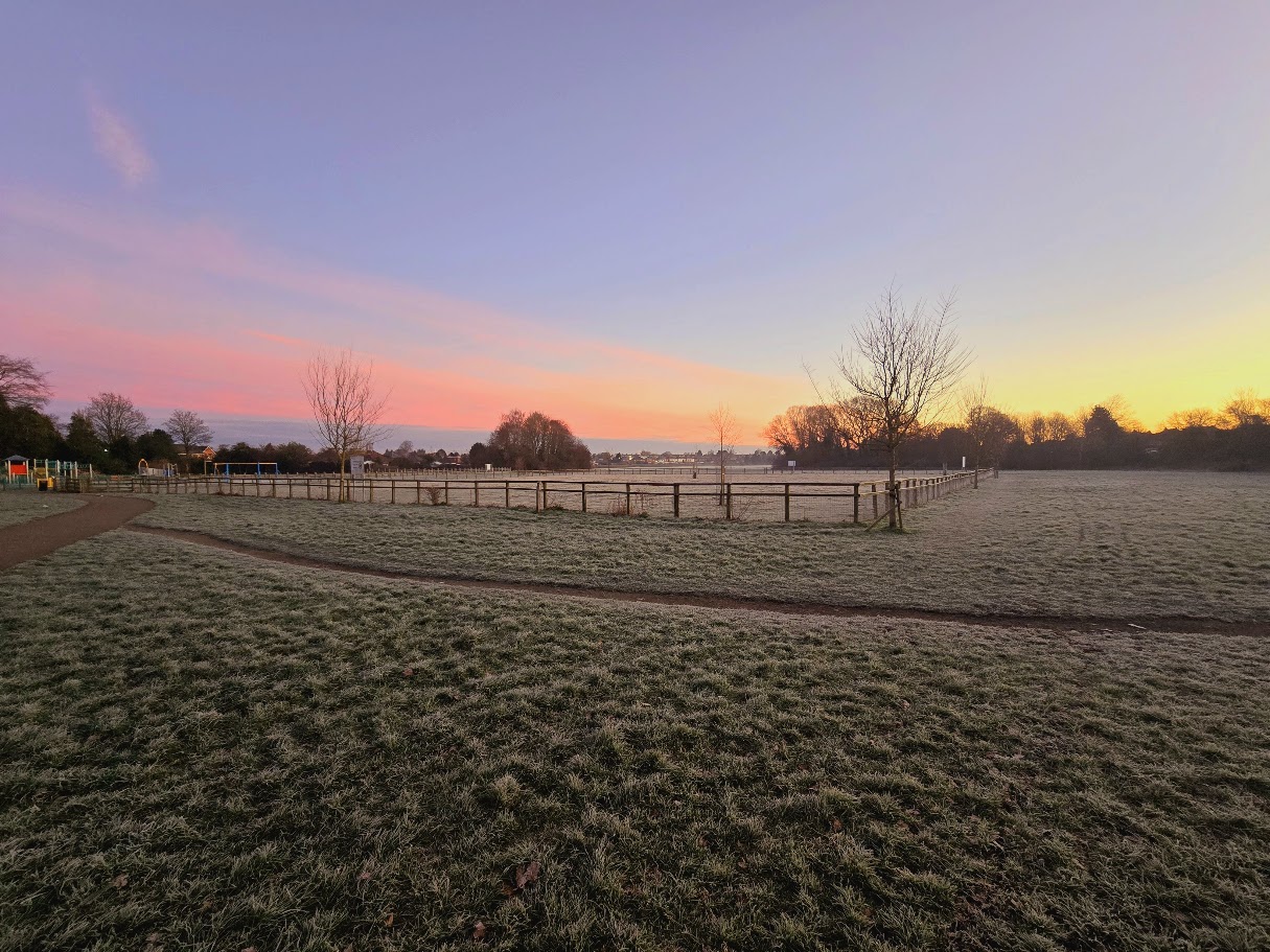 Frost-covered grass at dawn in Sheldon Country Park, with post-and-rail fencing around the farm fields, a children's playground visible to the left, and a vivid pink and orange sunrise sky above