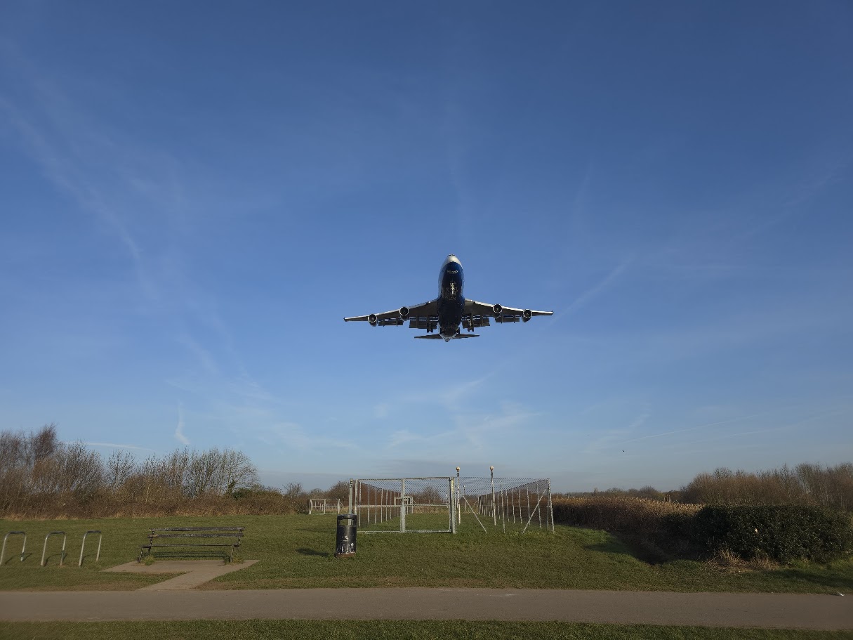 A large passenger aircraft on final approach to Birmingham Airport passing directly overhead at low altitude, seen from the viewing area at Sheldon Country Park, with park benches and a footpath visible below