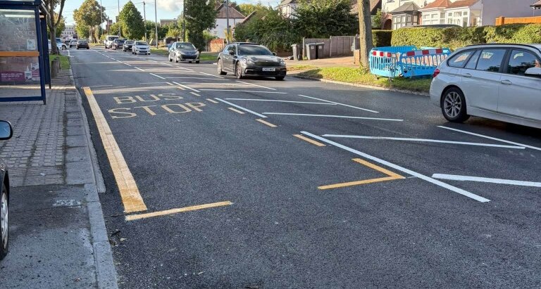 Freshly painted yellow BUS STOP markings on a newly resurfaced road in Sheldon, with lane dividers and roadwork barriers visible in the background
