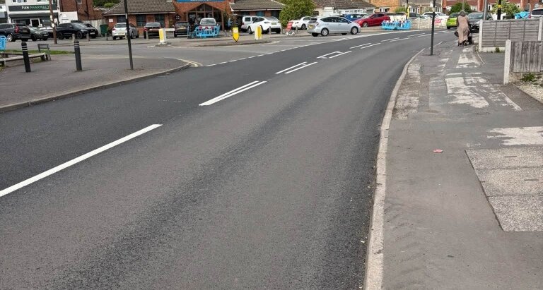 Newly resurfaced main road in Sheldon with fresh white lane markings on dark asphalt, with local shops and parked cars visible in the background